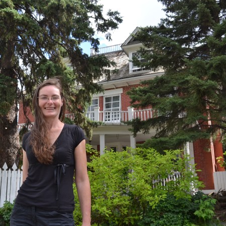Christy smiles on a sunny day. A large red brick house is behind her, but mostly blocked by trees.