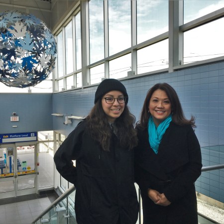 Lauren Crazybull and Sandy Pon stand inside the LRT station at Century Park, with a globe of blue leaves behind them.