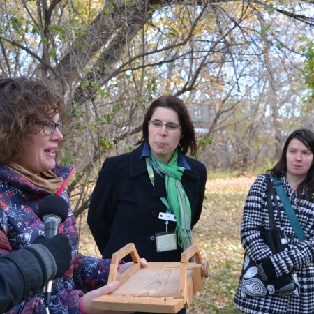 Heather Kerr speaks into a microphone while holding a small wooden model of John Walter's ferry, while Jade Dodd and Brittany Romano look on.
