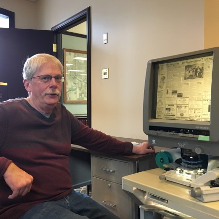 Glen Carlson is seated, looking at the camera, sitting in front of a microfilm reader