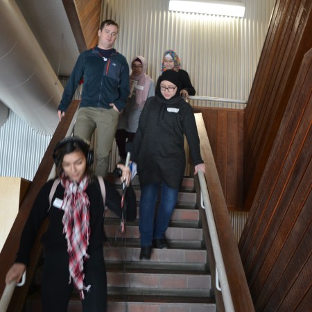 Rasha holds a microphone and wears headphones as she leads the way to Mike's building, with teacher Ian Potts behind her, as well as Laila, Mariam, and Nuha.