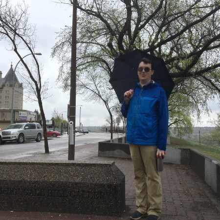 Allan stands holding an umbrella along a walkway, with the Hotel Macdonald in the background. To Allan's left, you can see the conrete plinth where the plaque/panel used to be.
