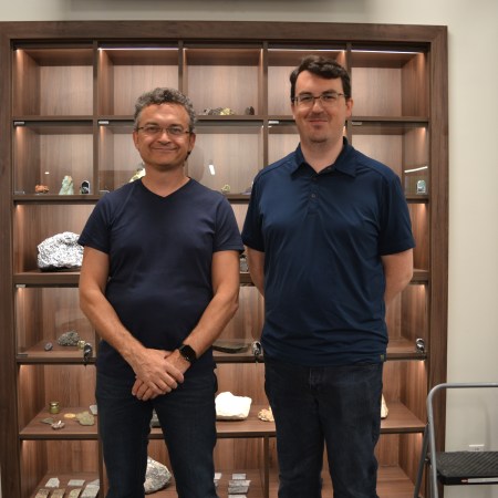 Jody Dahrouge and Allan stand in front of the wall of rocks at Dahrouge Geological Consulting. It's a glass case with about 30 display squares, each featuring a couple rocks and fossils.