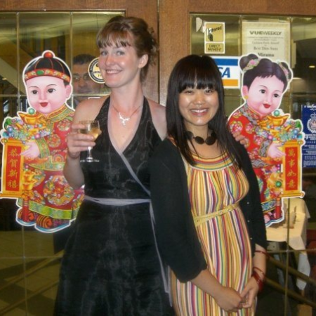Two women stand back to back in front of glass doors inside a restaurant, smiling