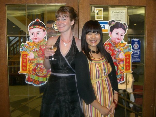 Two women stand back to back in front of glass doors inside a restaurant, smiling