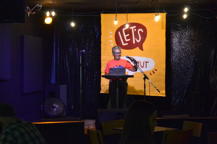 marilyn dumont smiles in front of a podium on stage at the aviary, with an orange t-shirt