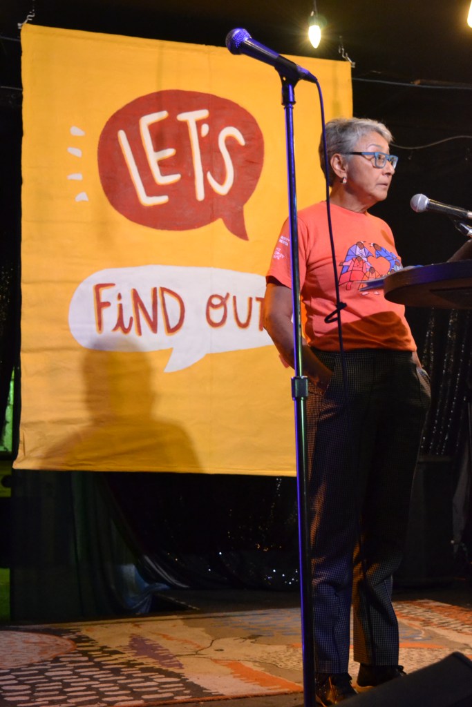 marilyn dumont reads poetry on stage in front of the let's find out banner