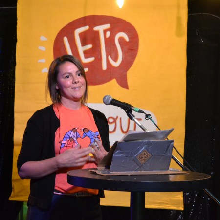 sarah de lano speaks in front of the let's find out banner, in an orange t-shirt