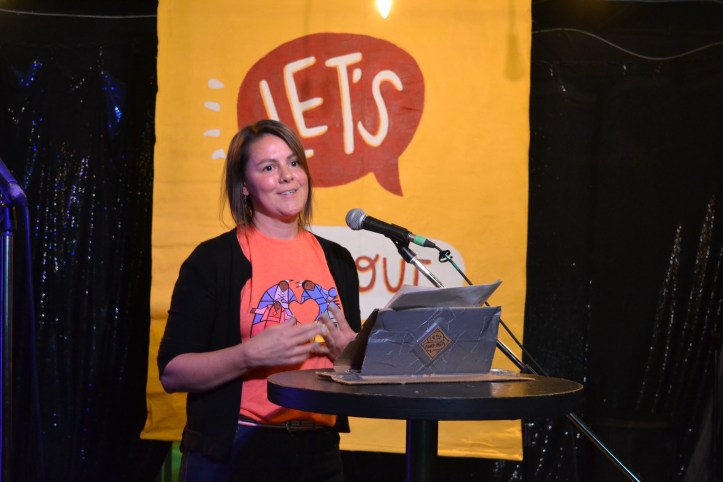 sarah de lano speaks in front of the let's find out banner, in an orange t-shirt