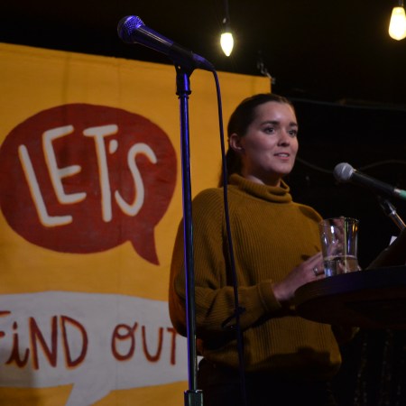 tara russell speaks at a podium in front of the let's find out banner, in a brown sweater