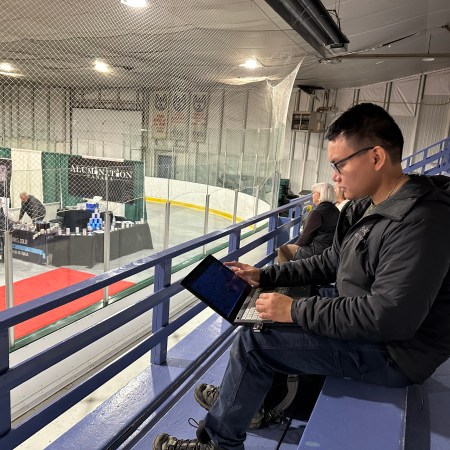 A man sits on a bench in the bleachers of an arena. To the right, a vendor is visible in the rink area.