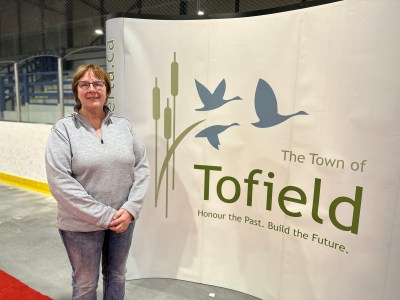 Vanita stands to the left of frame in front of a large sign with the Tofield logo: three birds flying past three cattails. 