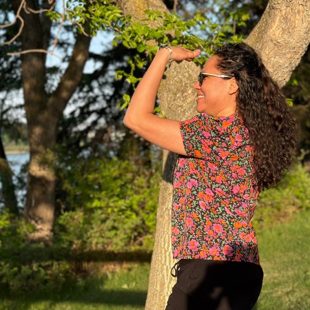 A woman with a colourful shirt and sunglasses stands in orange light in a park, staring off to the left towards a river.