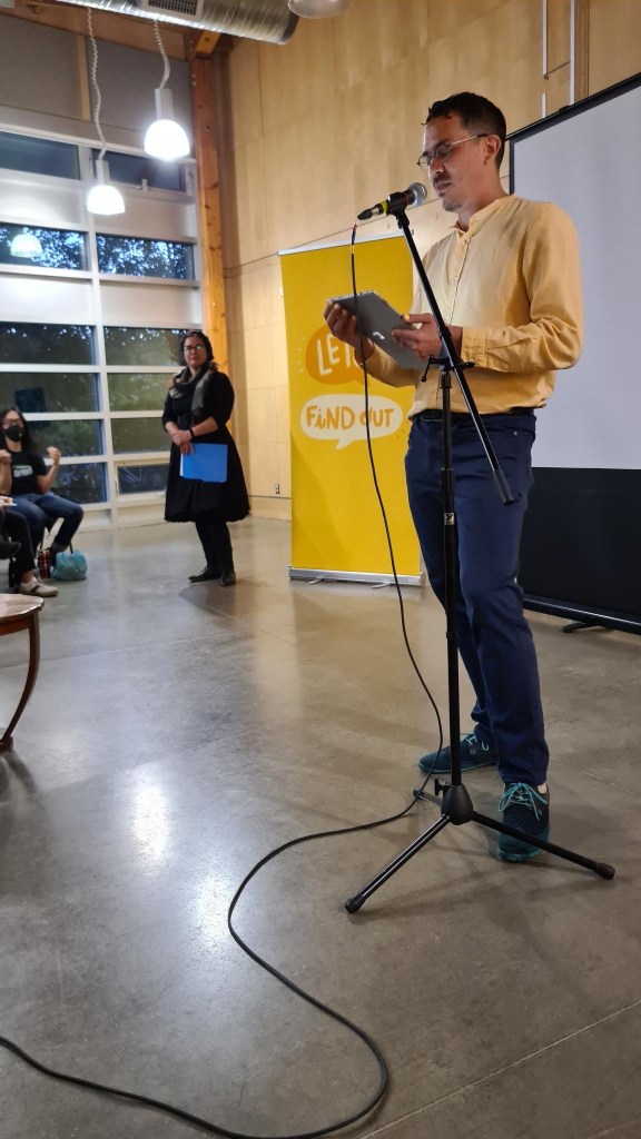 Chris Chang-Yen Phillips stands in front of a microphone, with the Let's Find Out banner and Lauren Markewicz behind him.