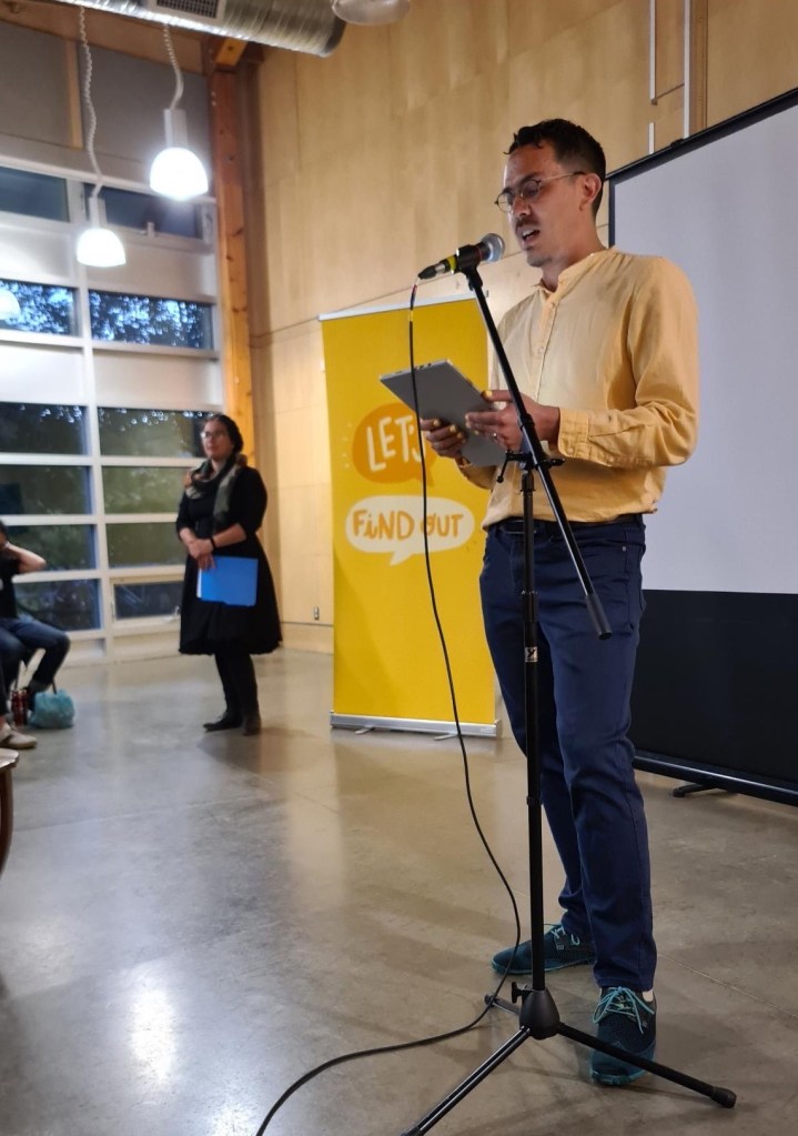 Chris Chang-Yen Phillips stands in front of a microphone, with the Let's Find Out banner and Lauren Markewicz behind him.