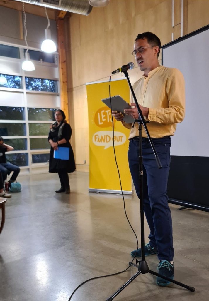 Chris Chang-Yen Phillips stands in front of a microphone, with the Let's Find Out banner and Lauren Markewicz behind him.