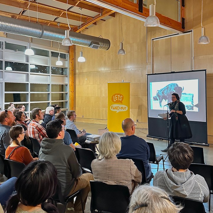 Speaker Lauren Markewicz talks at a microphone in front of a seated crowd. Behind her are the Let's Find Out banner and a projected image of bison.