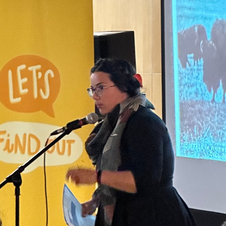 Speaker Lauren Markewicz talks at a microphone. Behind her are the Let's Find Out banner and a projected image of bison.