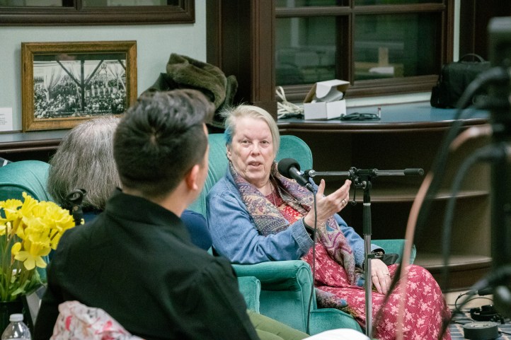 An older white woman sits in a comfortable chair, talking to two other people.