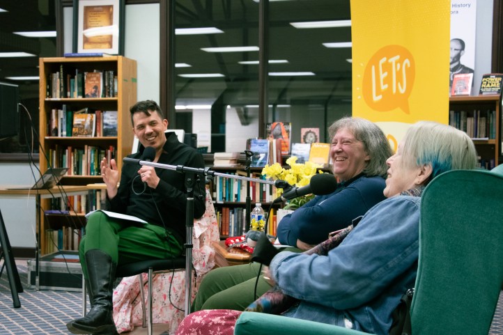 A mixed-race man in space boots interviews two older women in comfortable chairs.