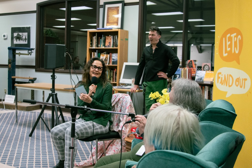 A stylish mixed-race woman sits in Chris' chair asking questions to the panelists