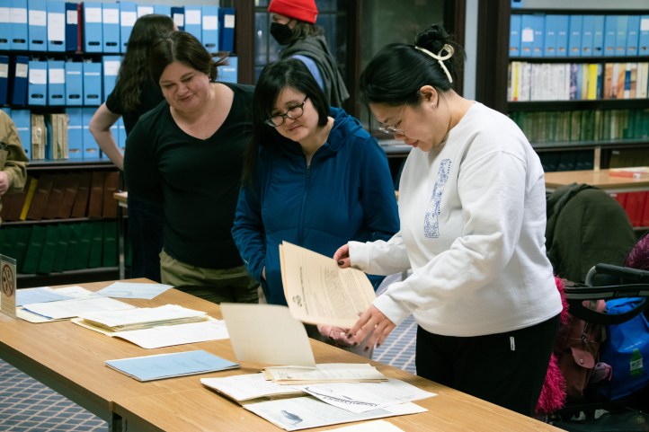 Three younger women look closely at papers spread out on a table at the archives